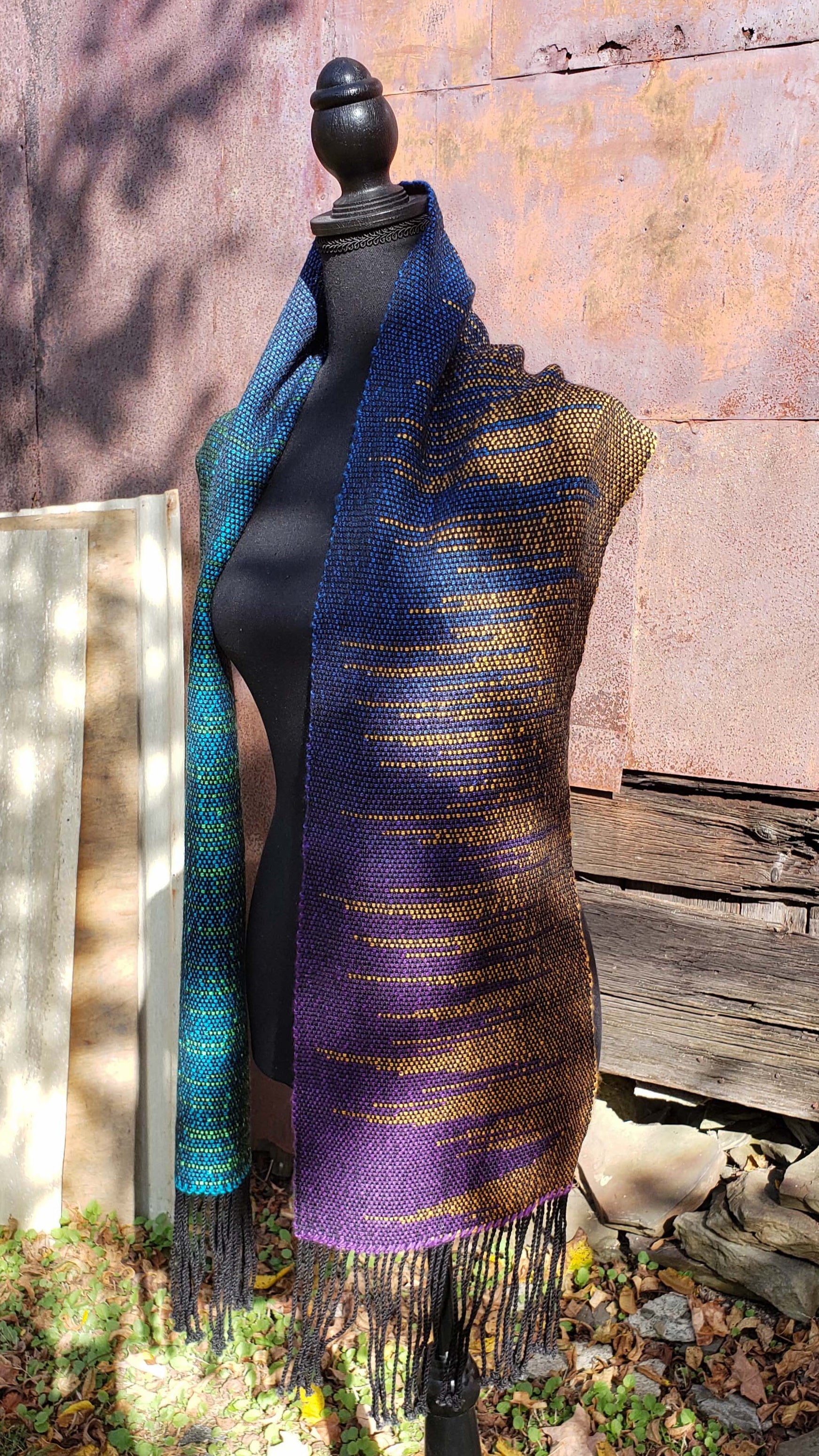 Front side view of a mannequin wearing a handwoven clasped weft scarf in gradient jewel tones with black fringe against the outside wall of a rustic building in an autumnal rural setting.