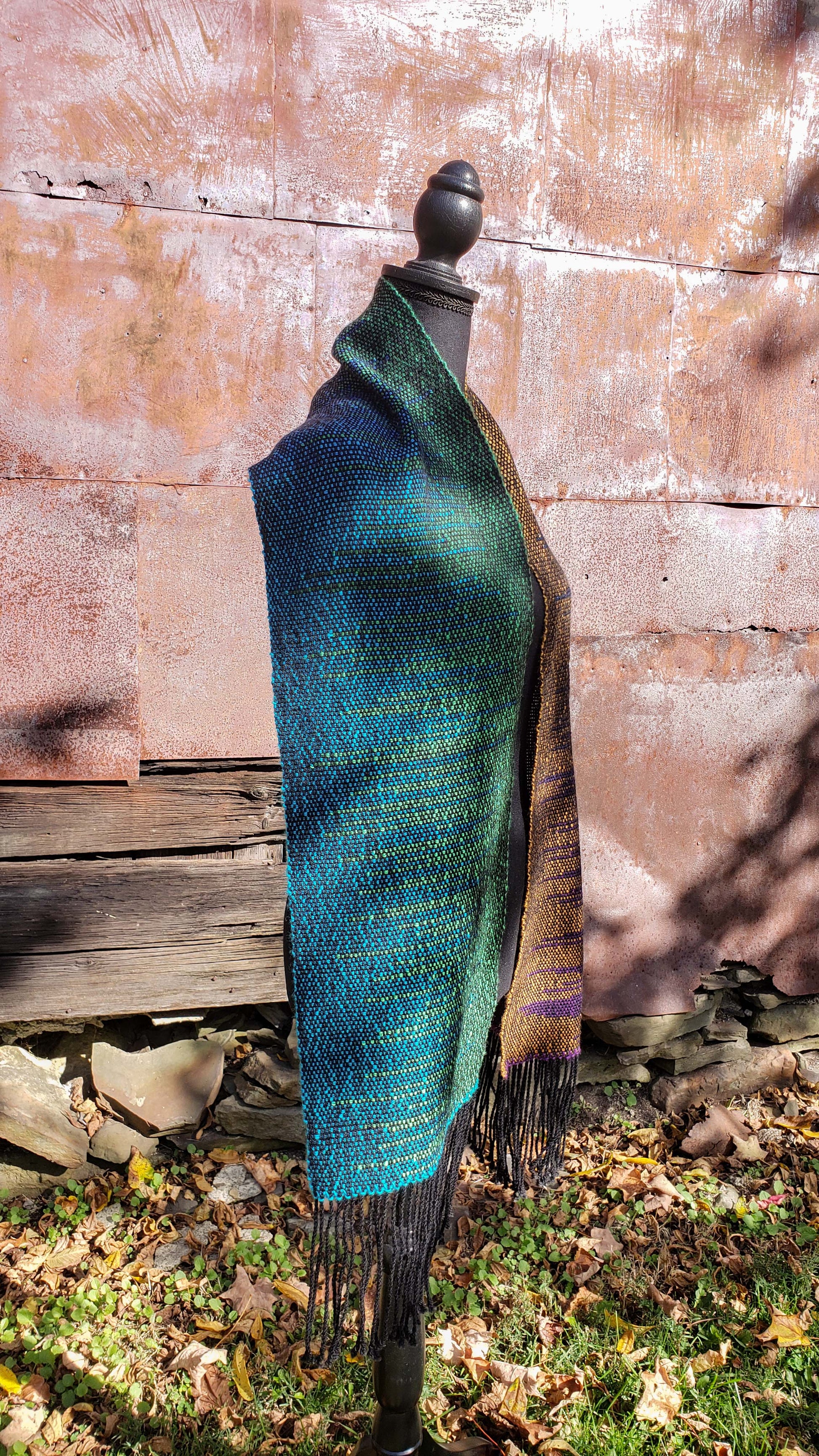Side view of a mannequin wearing a handwoven clasped weft scarf in gradient jewel tones with black fringe against the outside wall of a rustic building in an autumnal rural setting.