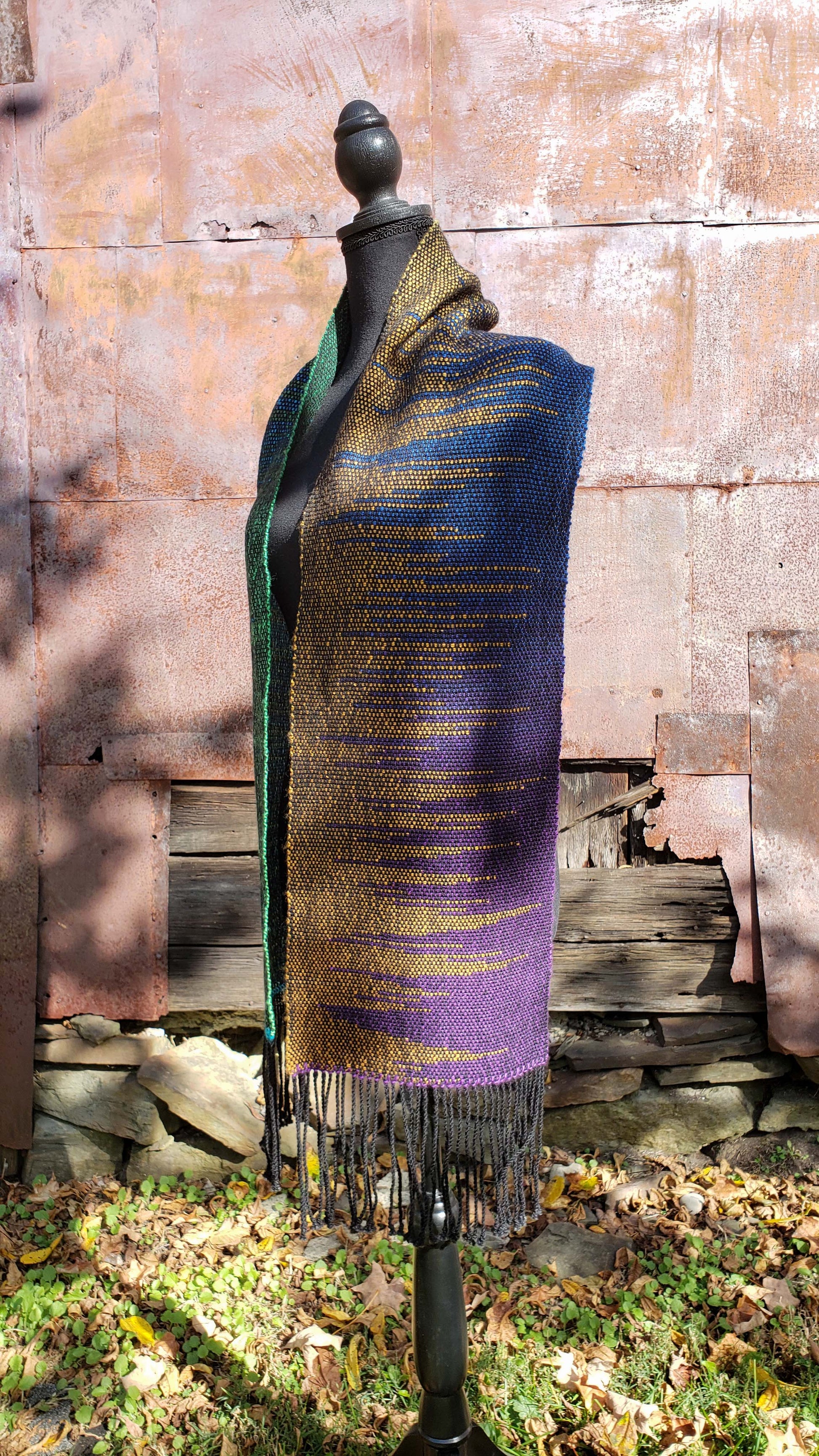 Side view of a mannequin wearing a handwoven clasped weft scarf in gradient jewel tones with black fringe against the outside wall of a rustic building in an autumnal rural setting.