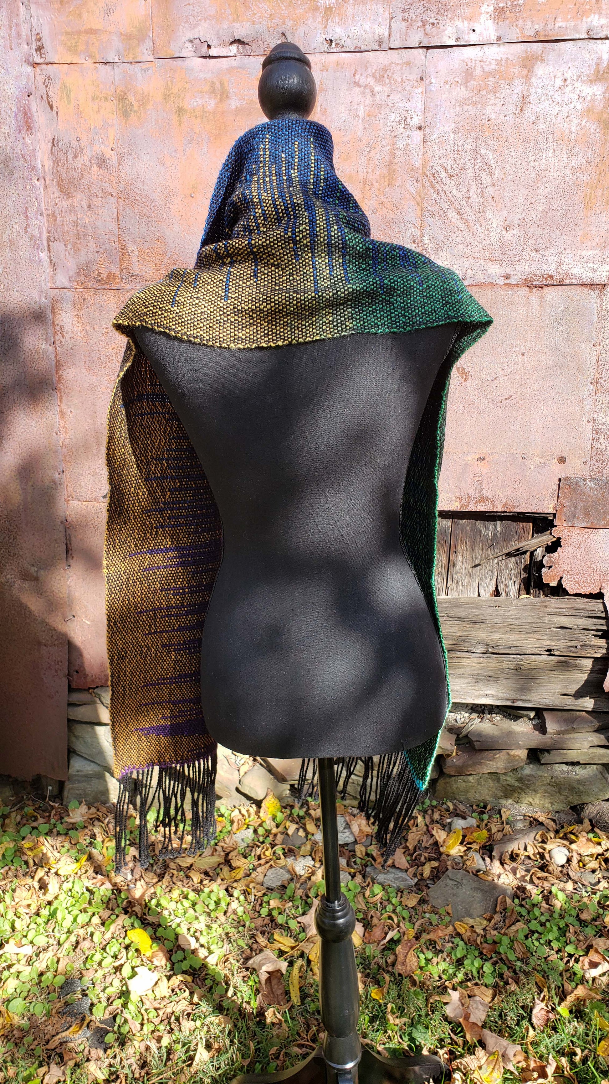 Rear view of a mannequin wearing a handwoven clasped weft scarf in gradient jewel tones with black fringe against the outside wall of a rustic building in an autumnal rural setting.