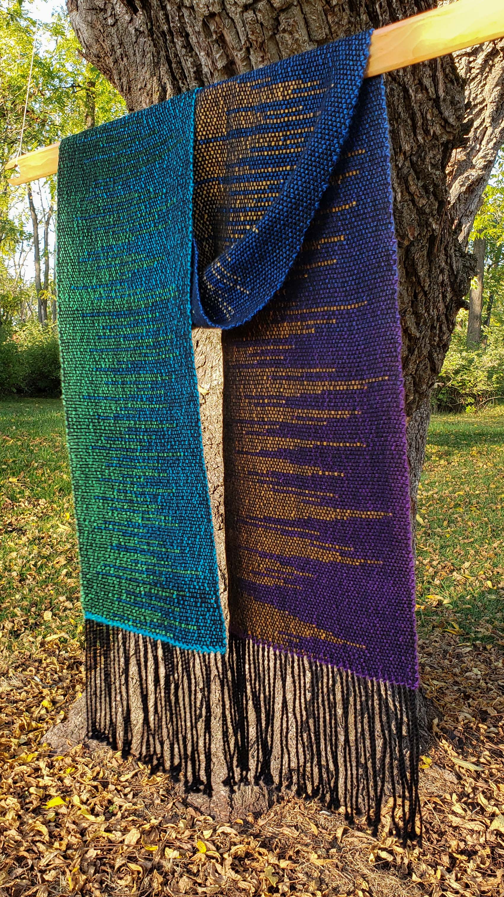 Colorful handwoven clasped weft scarf in jewel tones with black fringe draped over a weaving stick shuttle that is suspended from a tree in a natural autumnal setting