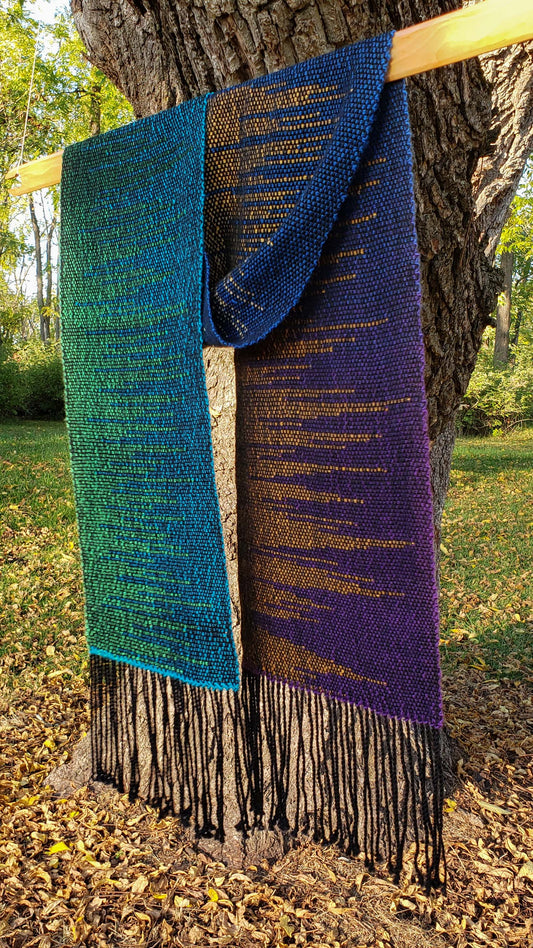 Colorful handwoven clasped weft scarf in jewel tones with black fringe draped over a weaving stick shuttle that is suspended from a tree in a natural autumnal setting