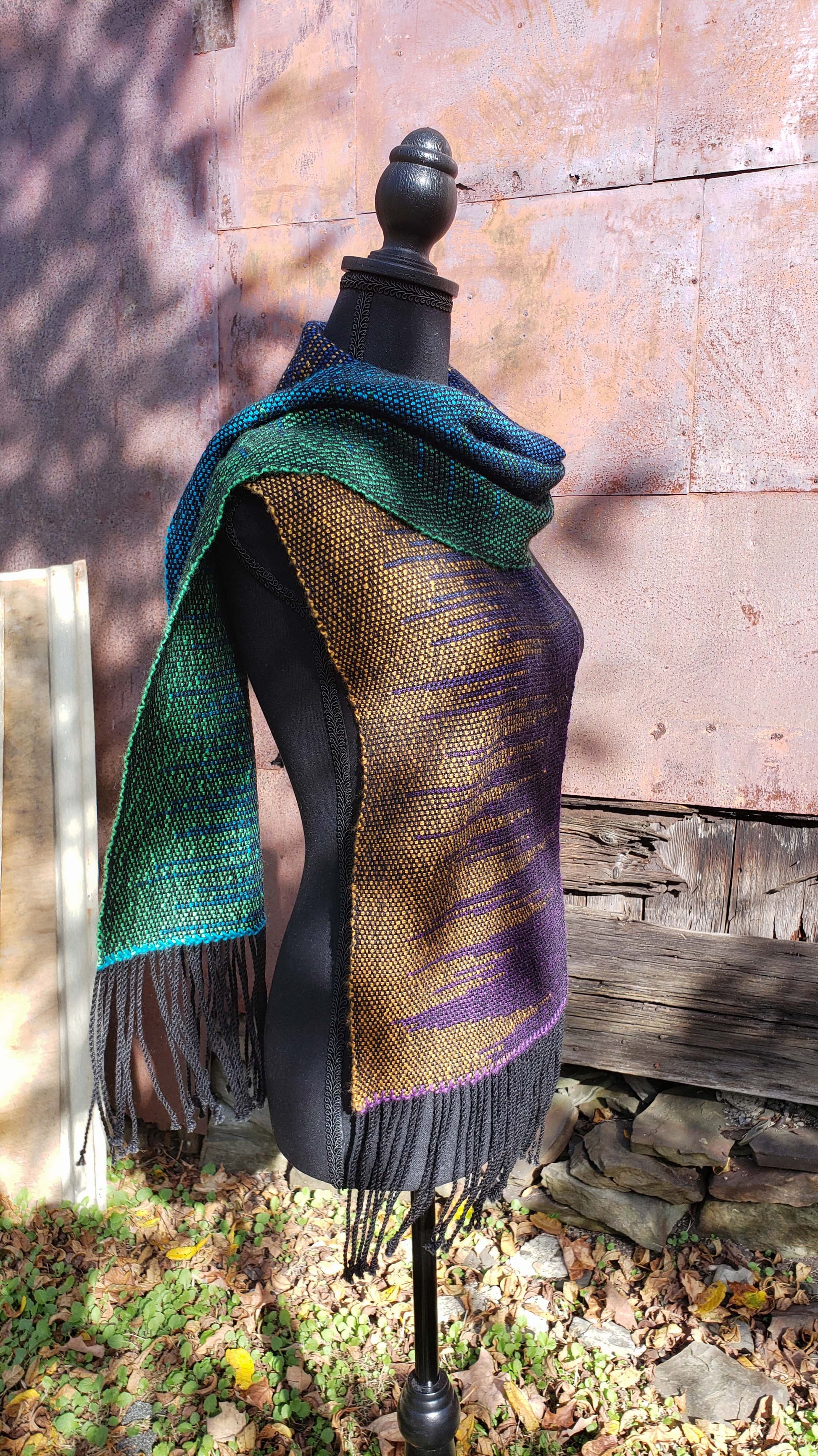 Side view of a mannequin wearing a handwoven clasped weft scarf in gradient jewel tones with black fringe against the outside wall of a rustic building in an autumnal rural setting.