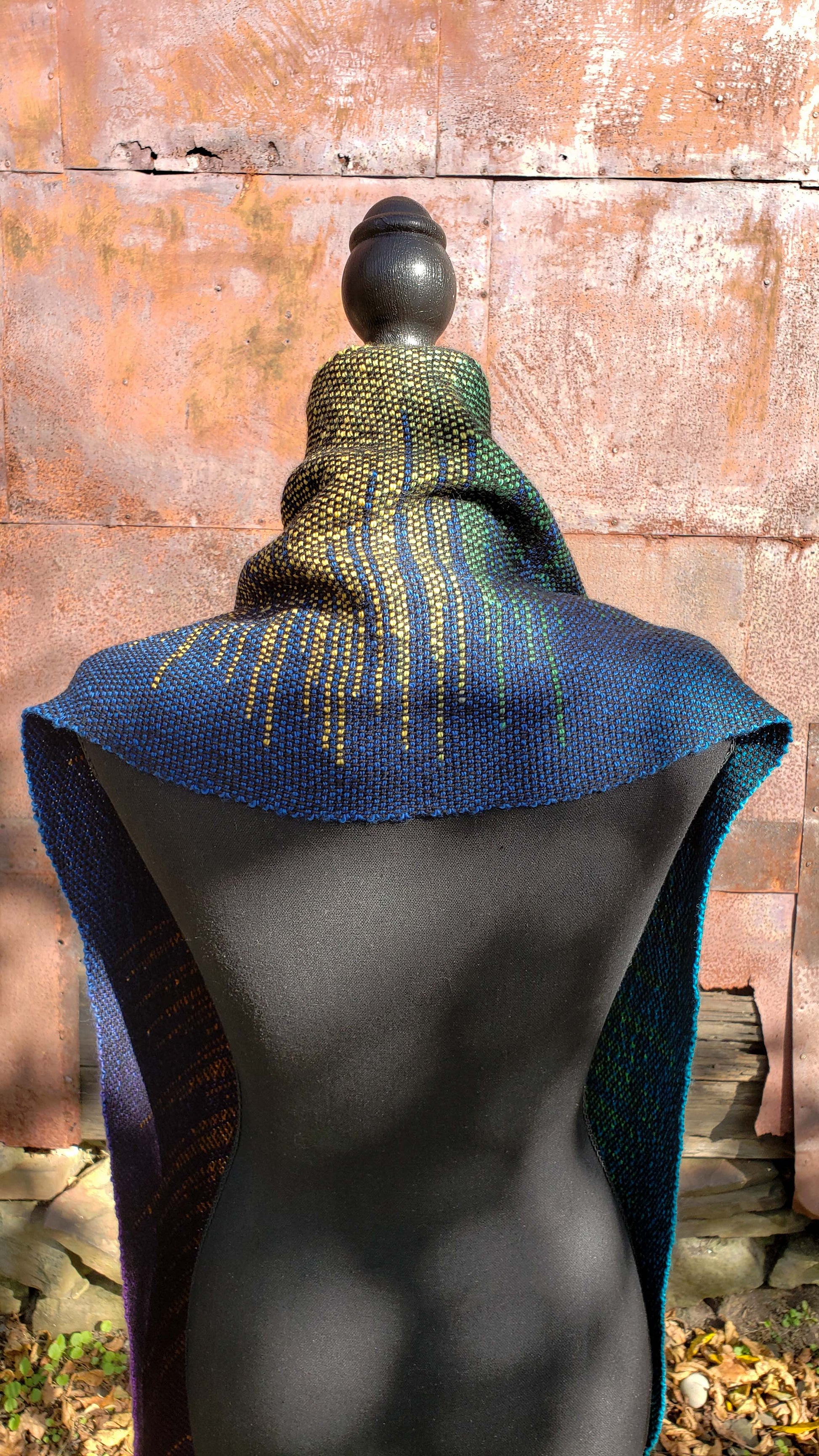 Rear view of a mannequin wearing a handwoven clasped weft scarf in gradient jewel tones with black fringe against the outside wall of a rustic building in an autumnal rural setting.