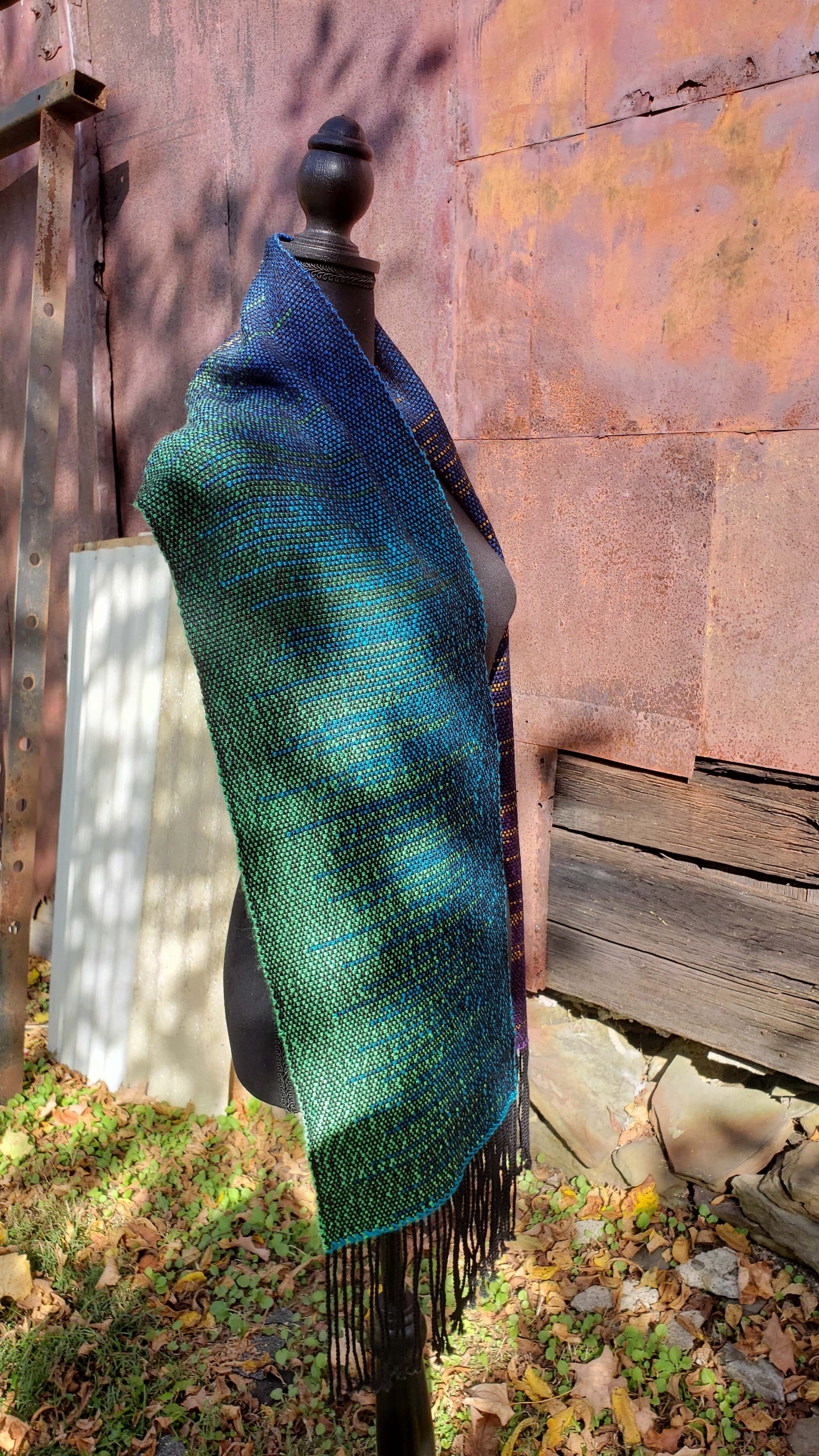 Side view of a mannequin wearing a handwoven clasped weft scarf in gradient jewel tones with black fringe against the outside wall of a rustic building in an autumnal rural setting.