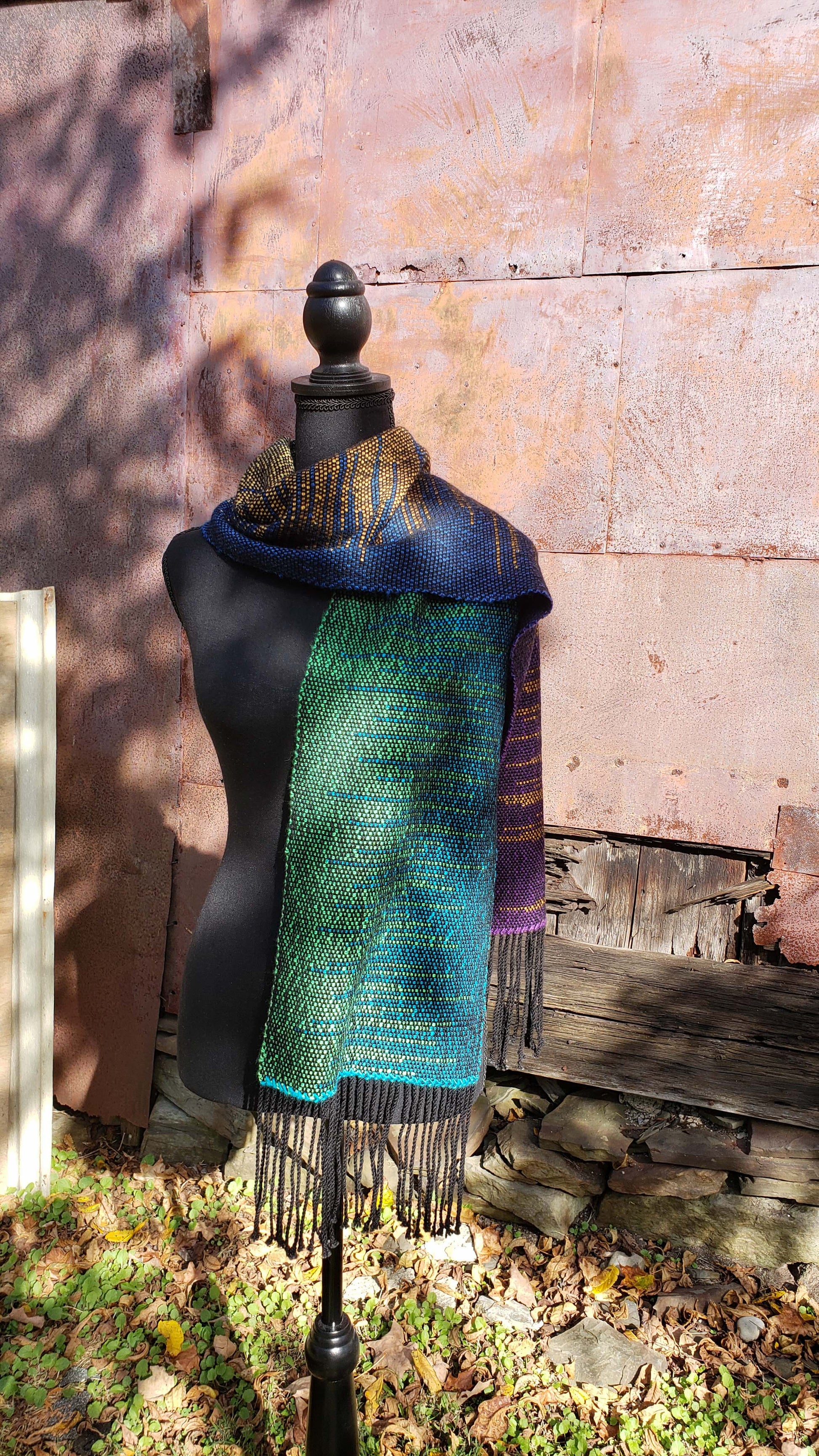 Front view of a mannequin wearing a handwoven clasped weft scarf in gradient jewel tones with black fringe against the outside wall of a rustic building in an autumnal rural setting.