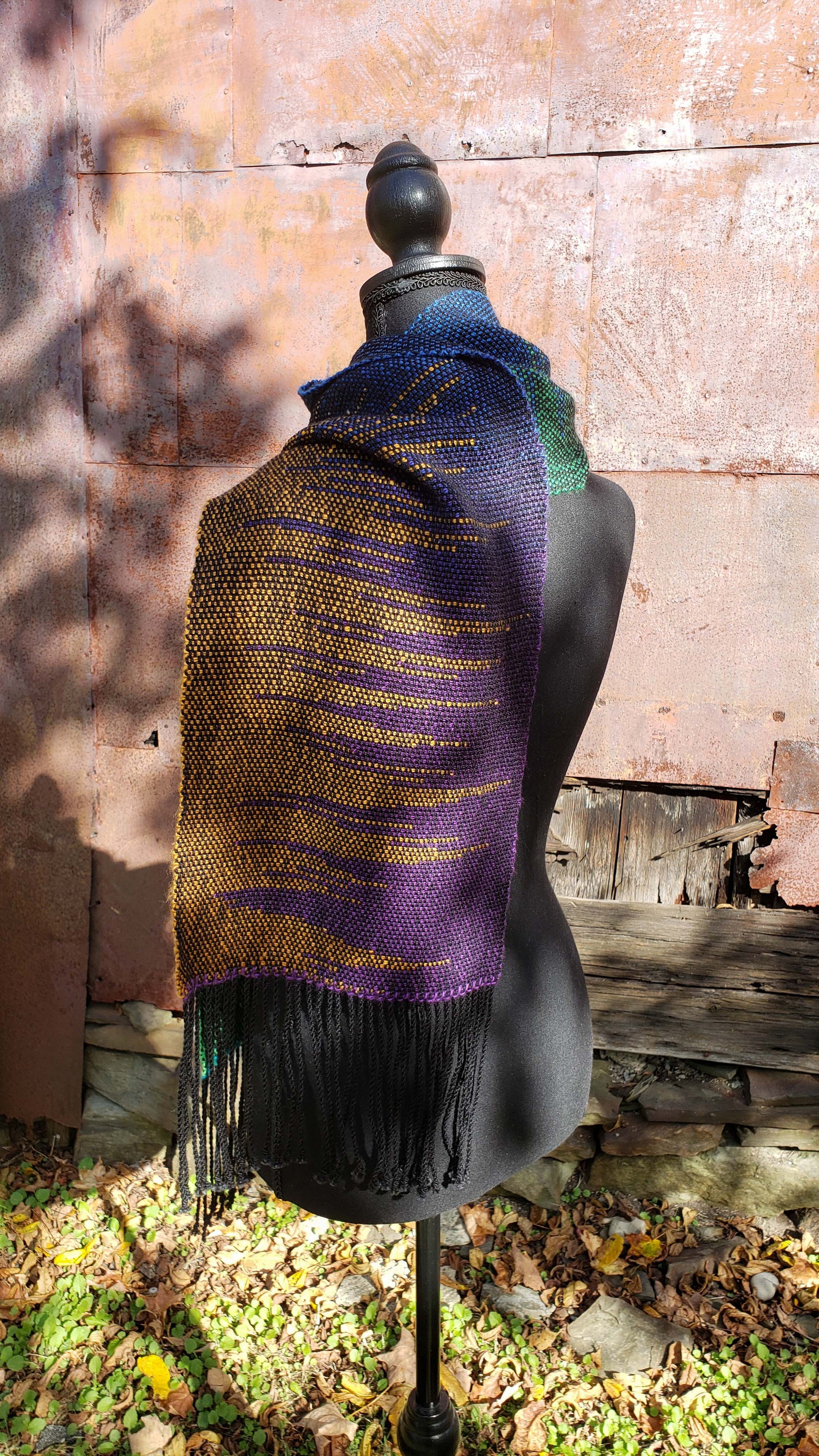 Rear side view of a mannequin wearing a handwoven clasped weft scarf in gradient jewel tones with black fringe against the outside wall of a rustic building in an autumnal rural setting.
