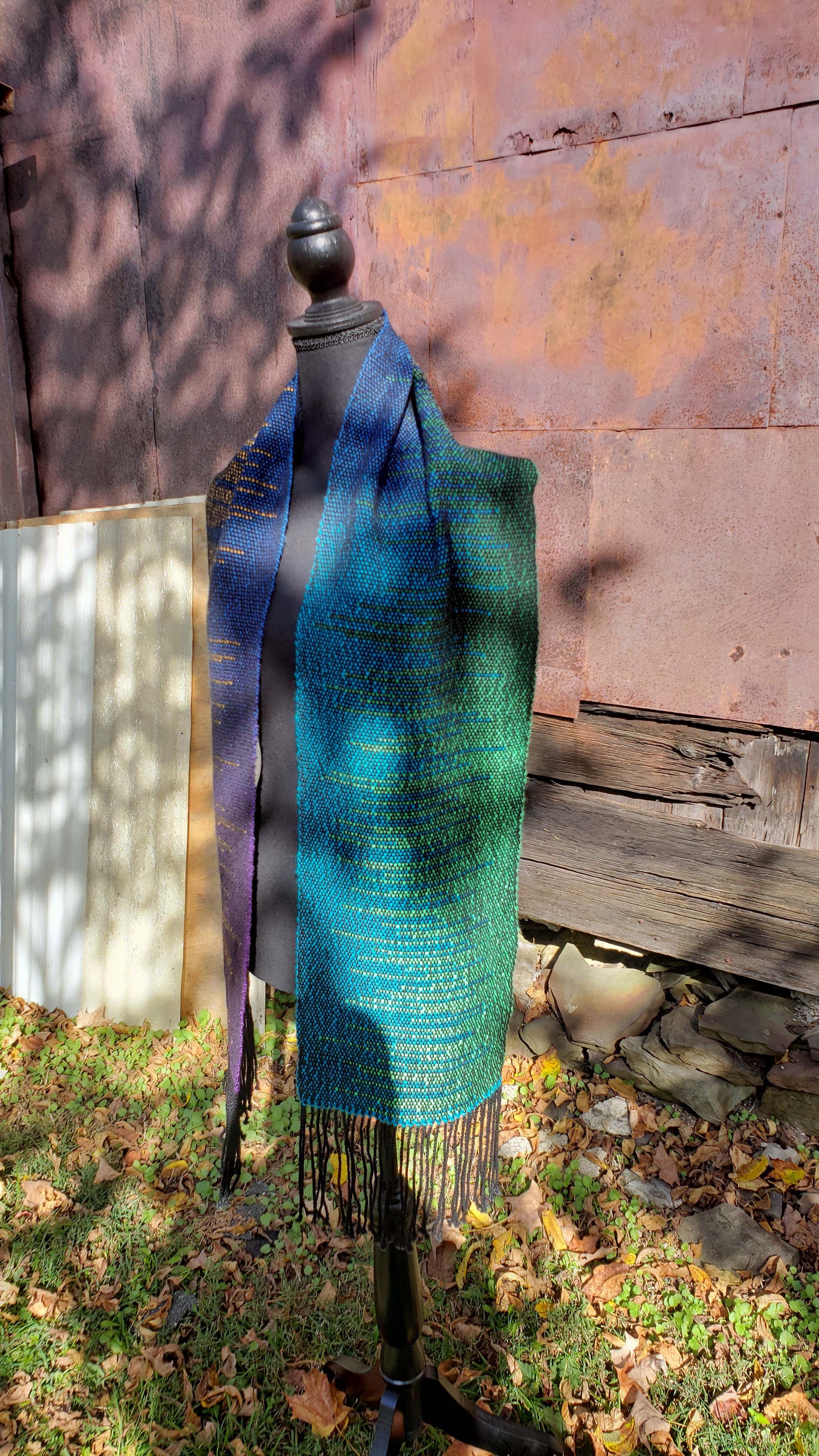 Front view of a mannequin wearing a handwoven clasped weft scarf in gradient jewel tones with black fringe against the outside wall of a rustic building in an autumnal rural setting.