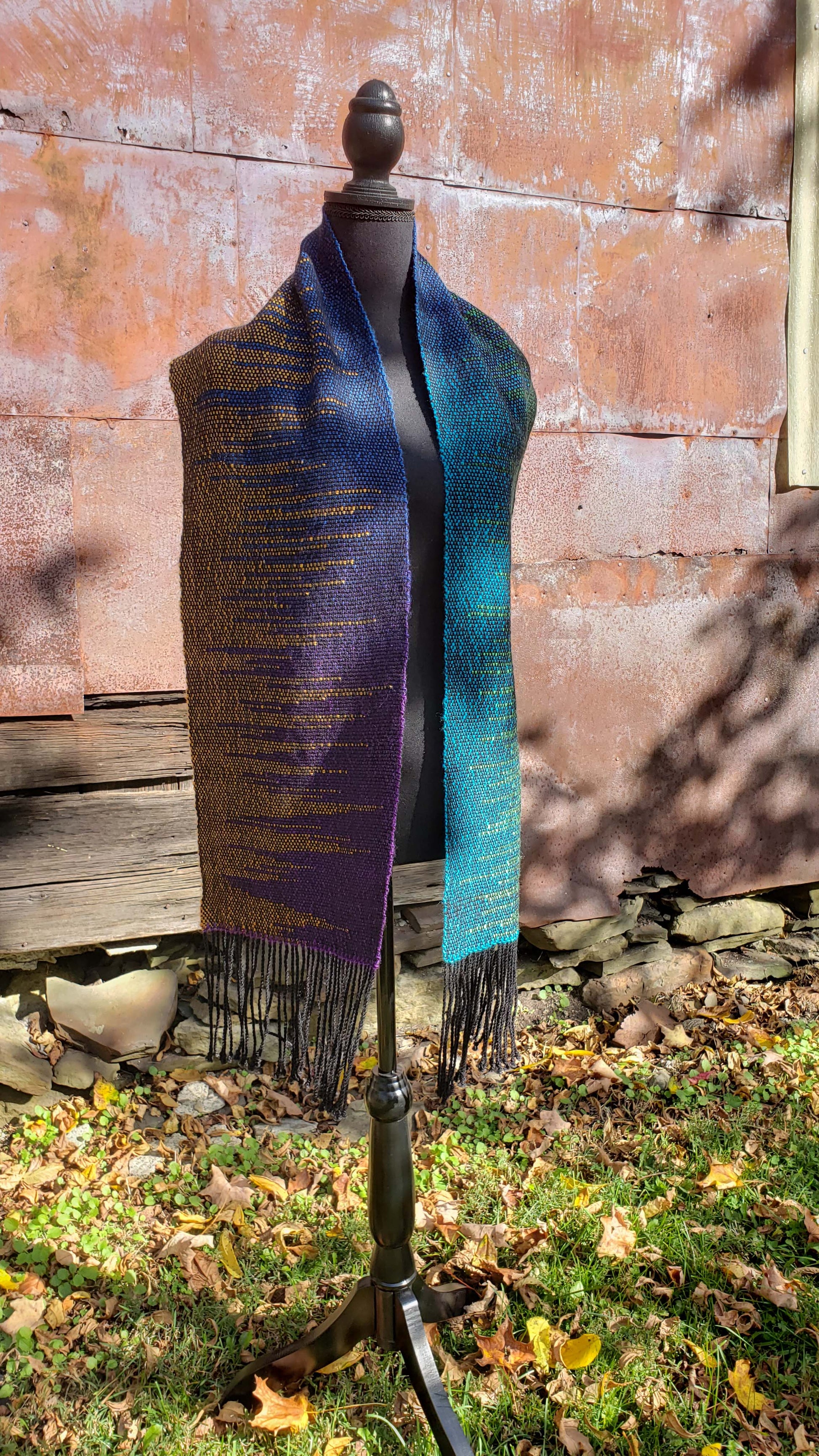 Front view of a mannequin wearing a handwoven clasped weft scarf in gradient jewel tones with black fringe against the outside wall of a rustic building in an autumnal rural setting.