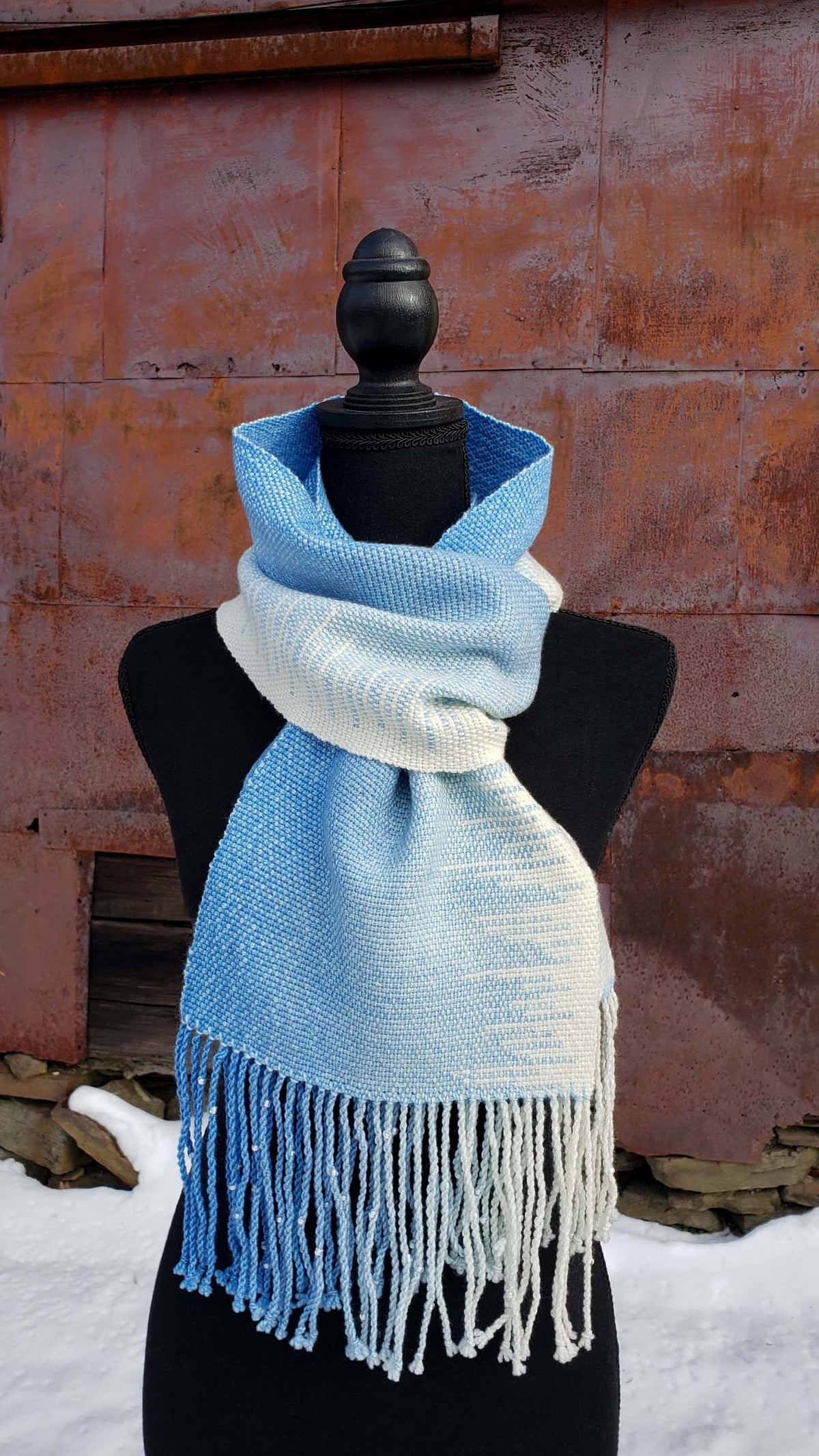 Blue and white handwoven scarf on a mannequin against a rustic exterior wall with a layer of snow on the ground.