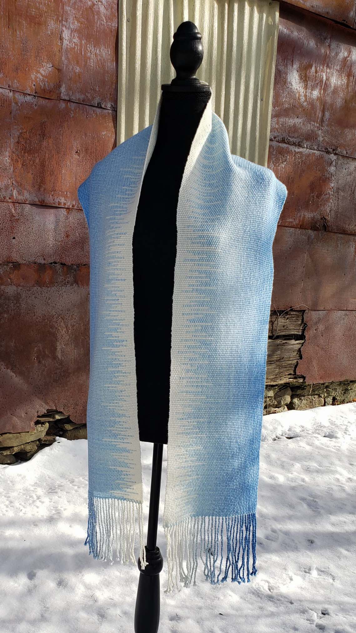 Blue and white handwoven scarf on a mannequin against a rustic exterior wall with a layer of snow on the ground.