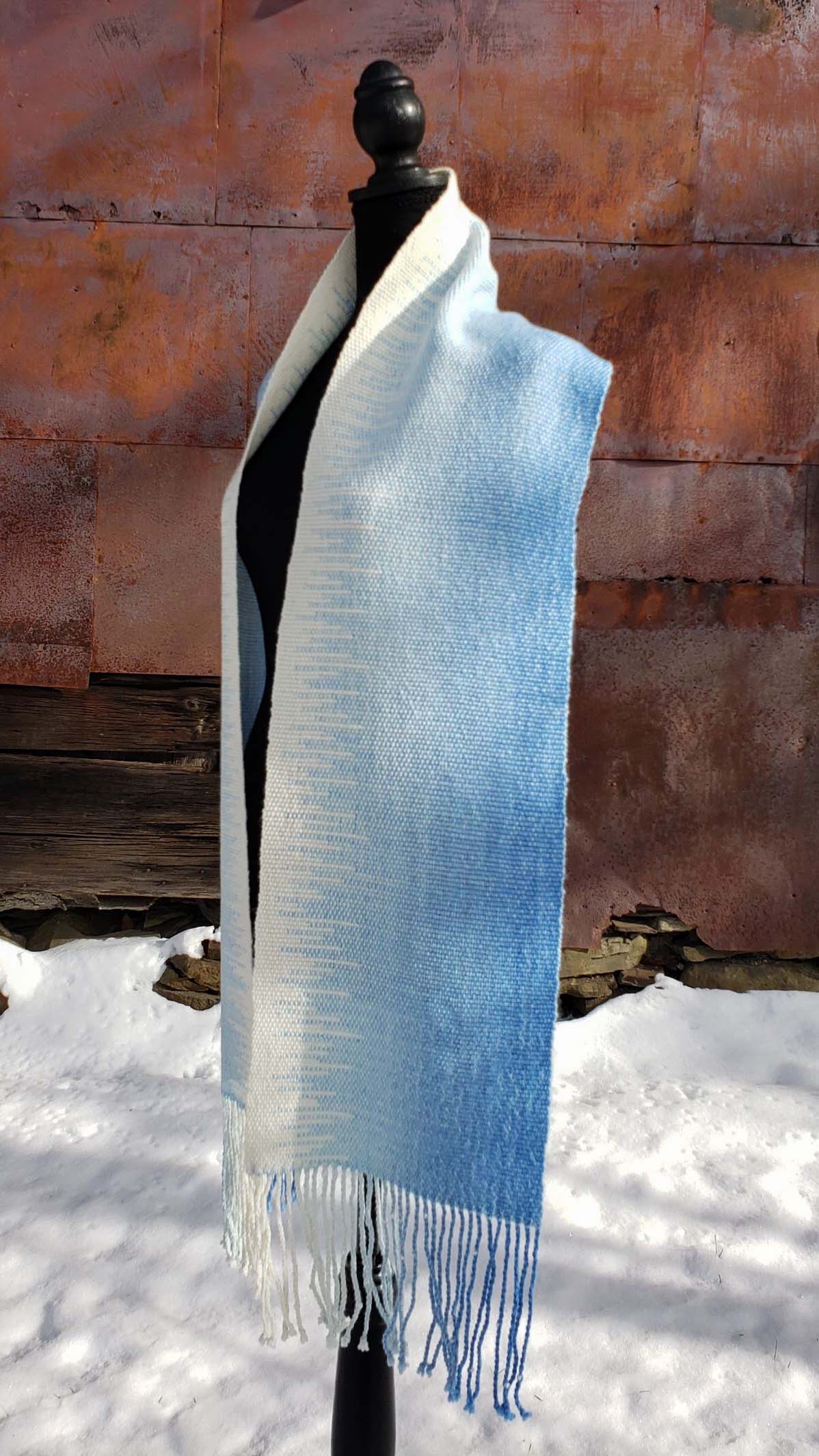 Blue and white handwoven scarf on a mannequin against a rustic exterior wall with a layer of snow on the ground.