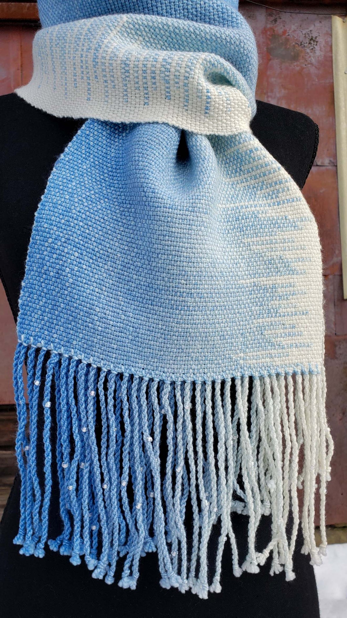 Blue and white handwoven scarf on a mannequin against a rustic exterior wall with a layer of snow on the ground.
