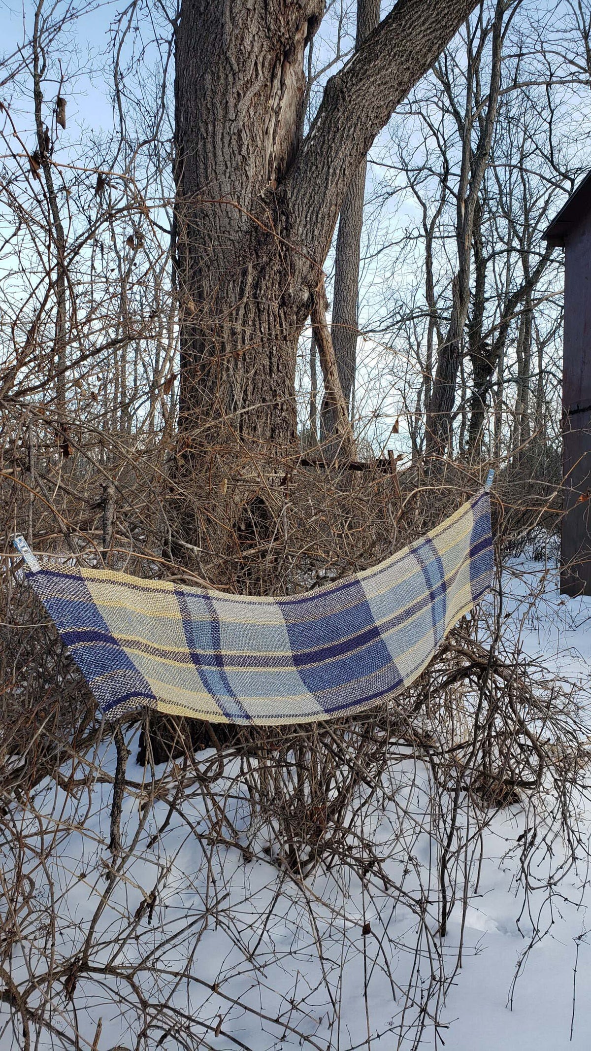 Plaid handwoven table runner draped over branches in a snowy landscape with bare trees.