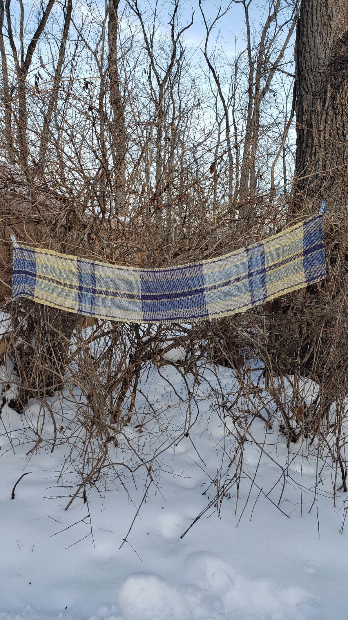 Handwoven plaid table runner draped across branches in a snowy wooded background.