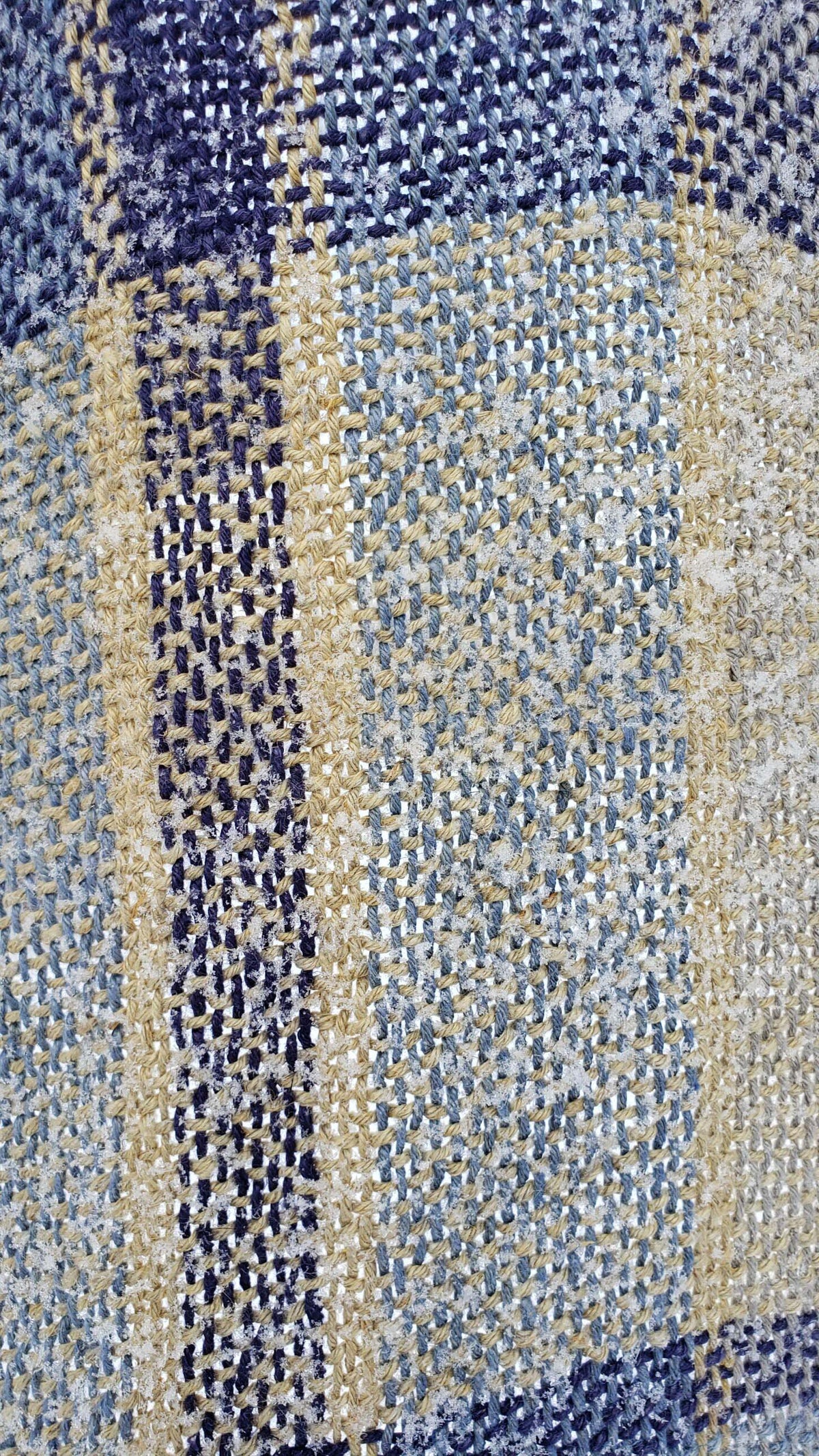 Close-up of a snow dusted handwoven fabric with blue, yellow, and gray plaid pattern.