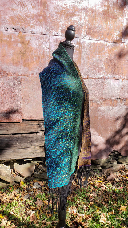 Side view of a mannequin wearing a handwoven clasped weft scarf in gradient jewel tones with black fringe against the outside wall of a rustic building in an autumnal rural setting.