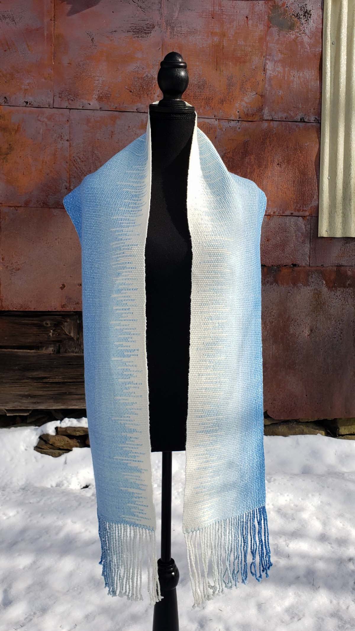 Blue and white handwoven scarf on a mannequin against a rustic exterior wall with a layer of snow on the ground.