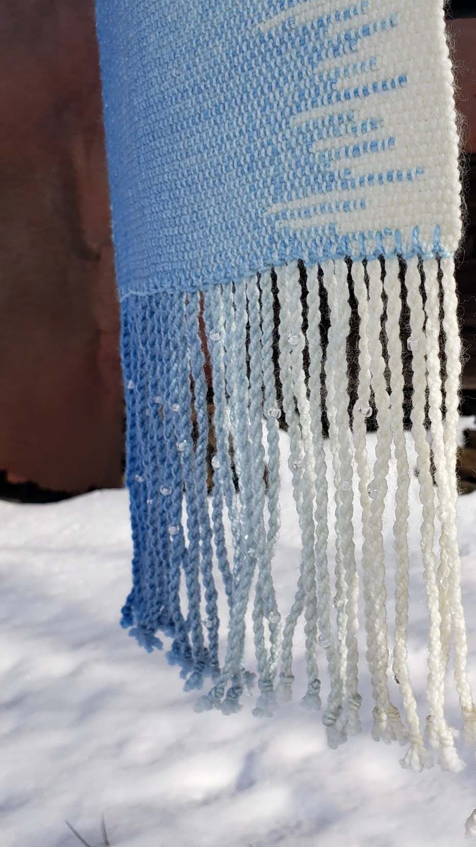 Closeup of a blue and white handwoven scarf with beaded twisted fringe against a rustic exterior wall with a layer of snow on the ground.