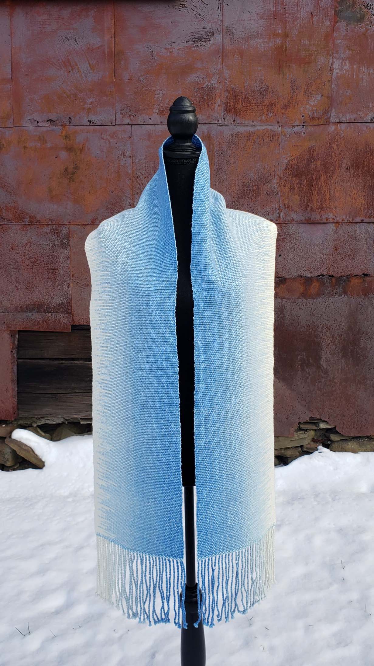 Blue and white handwoven scarf on a mannequin against a rustic exterior wall with a layer of snow on the ground.