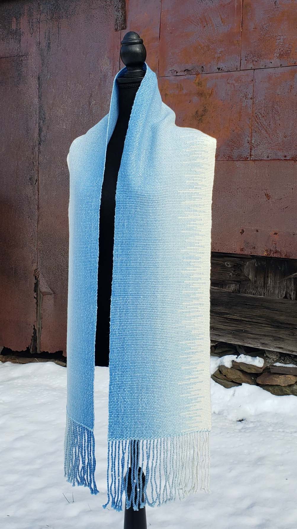 Blue and white handwoven scarf on a mannequin against a rustic exterior wall with a layer of snow on the ground.