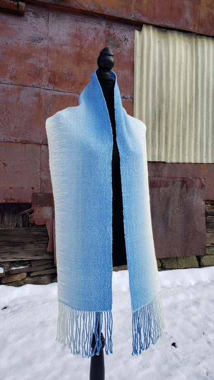 Blue and white handwoven scarf on a mannequin against a rustic exterior wall with a layer of snow on the ground.