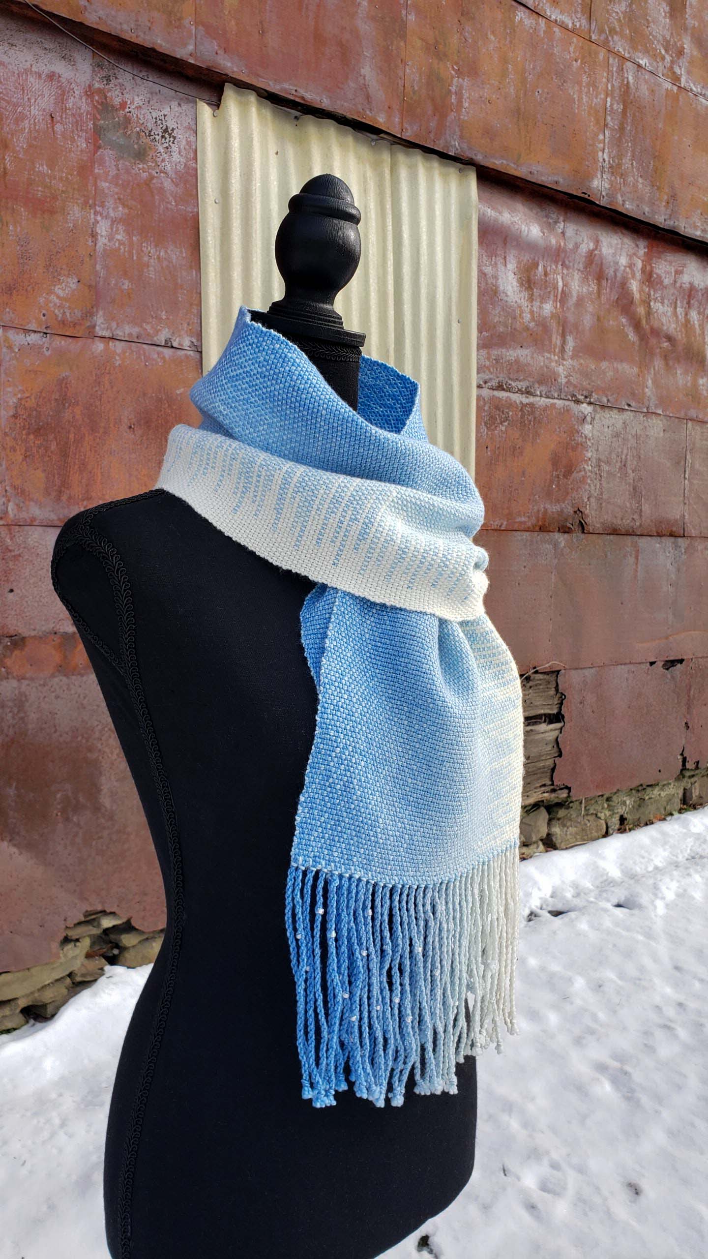 Blue and white handwoven scarf on a mannequin against a rustic exterior wall with a layer of snow on the ground.
