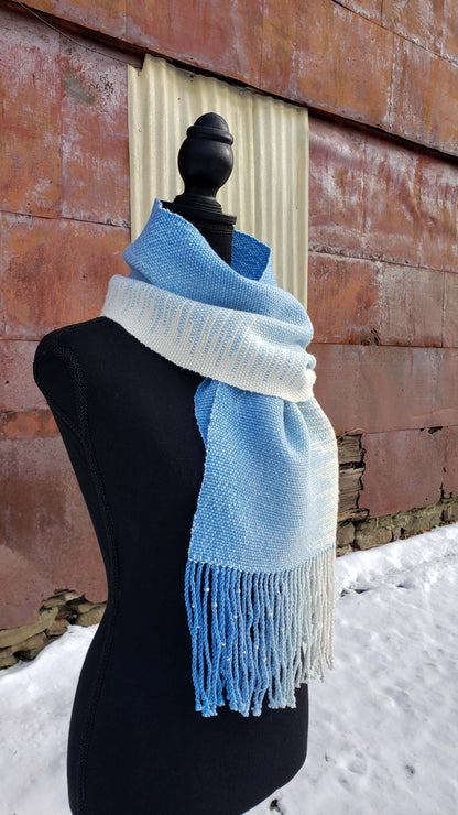Blue and white handwoven scarf on a mannequin against a rustic exterior wall with a layer of snow on the ground.
