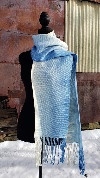 Blue and white handwoven scarf on a mannequin against a rustic exterior wall with a layer of snow on the ground.