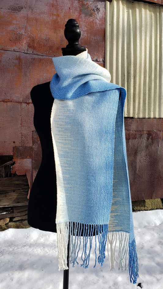 Blue and white handwoven scarf on a mannequin against a rustic exterior wall with a layer of snow on the ground.