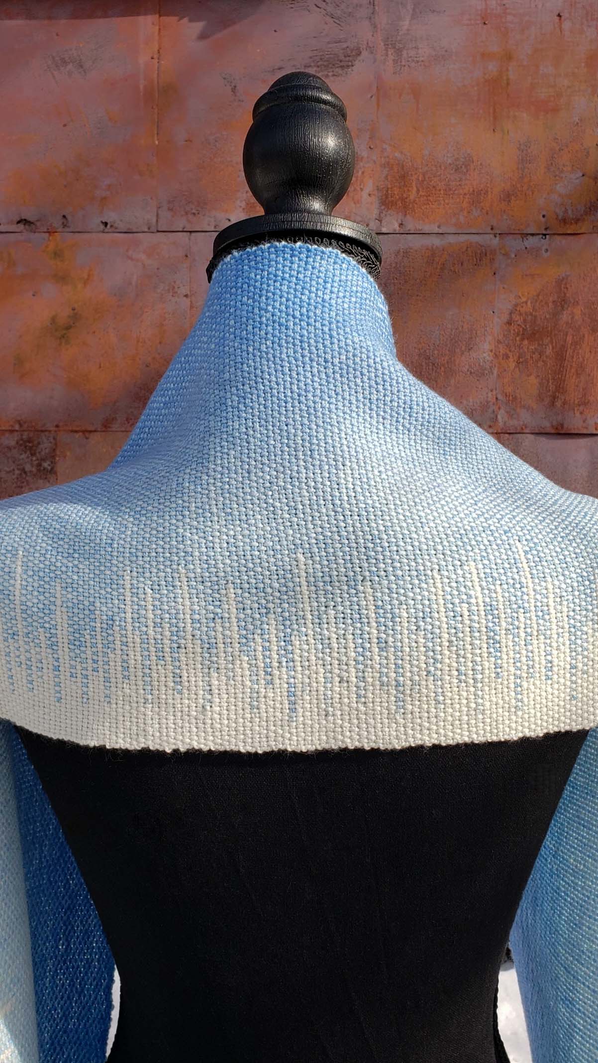 Blue and white handwoven scarf on a mannequin against a rustic exterior wall with a layer of snow on the ground.