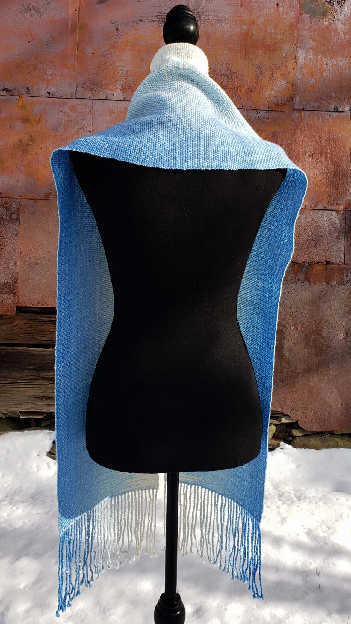 Blue and white handwoven scarf on a mannequin against a rustic exterior wall with a layer of snow on the ground.