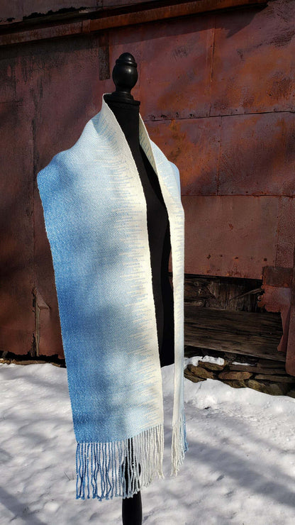 Blue and white handwoven scarf on a mannequin against a rustic exterior wall with a layer of snow on the ground.