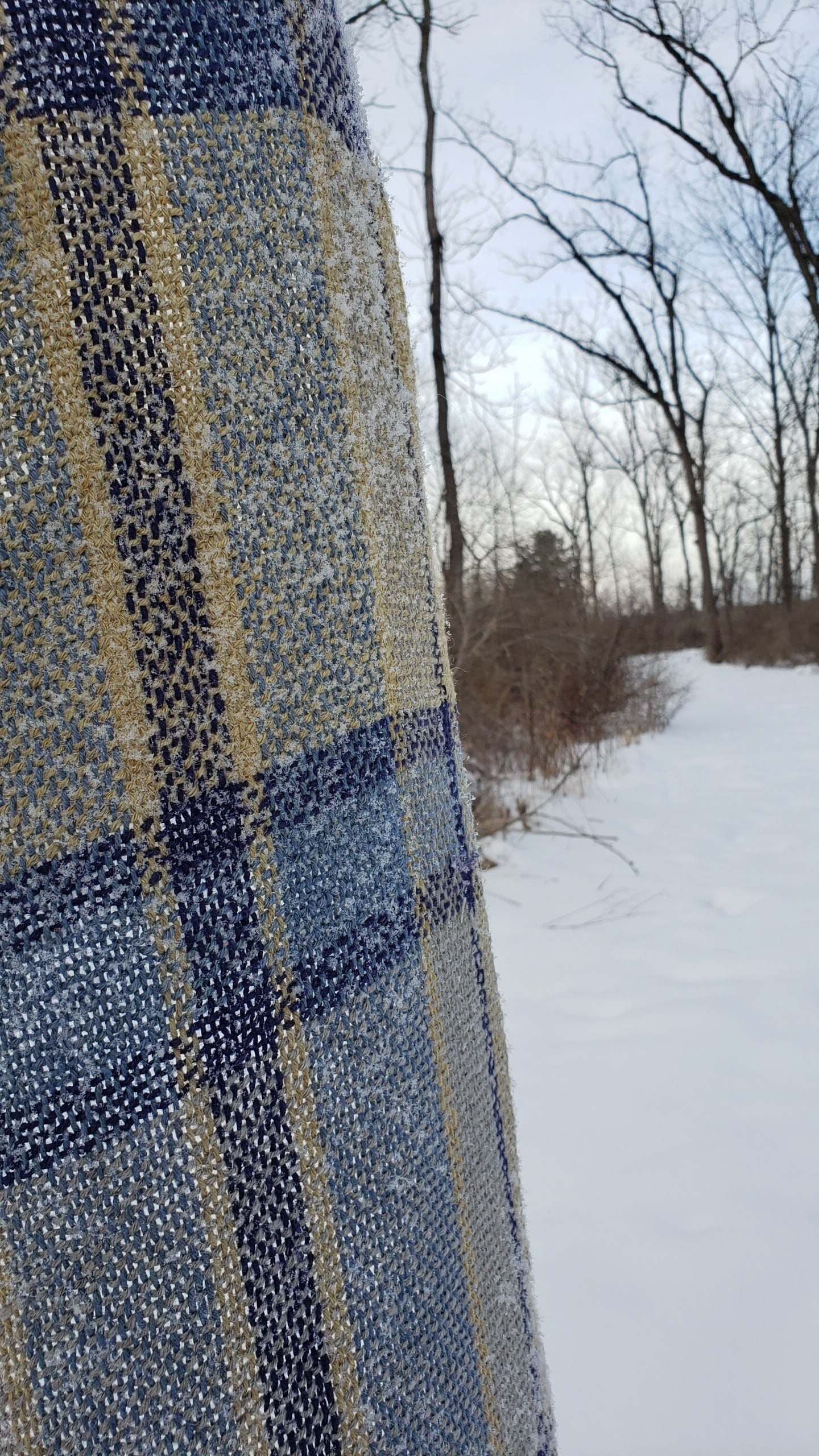 Handwoven plaid table runner with a dusting of snow against a snowy background with bare trees.
