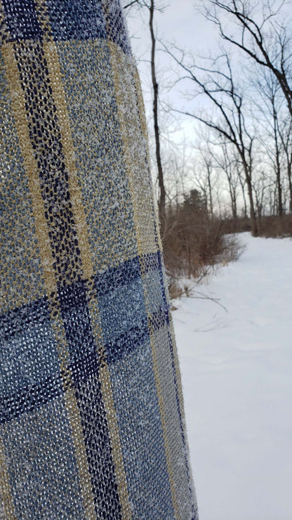 Handwoven plaid table runner with a dusting of snow against a snowy background with bare trees.
