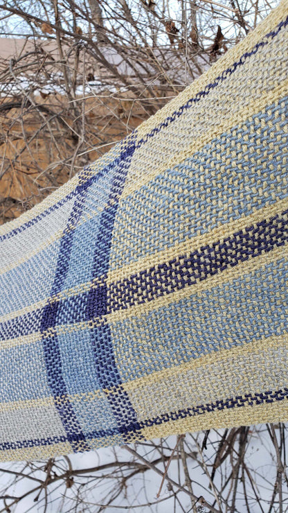 Closeup of a handwoven table runner with blue, grey and yellow plaid pattern draped over branches with bare trees and snow in the background.