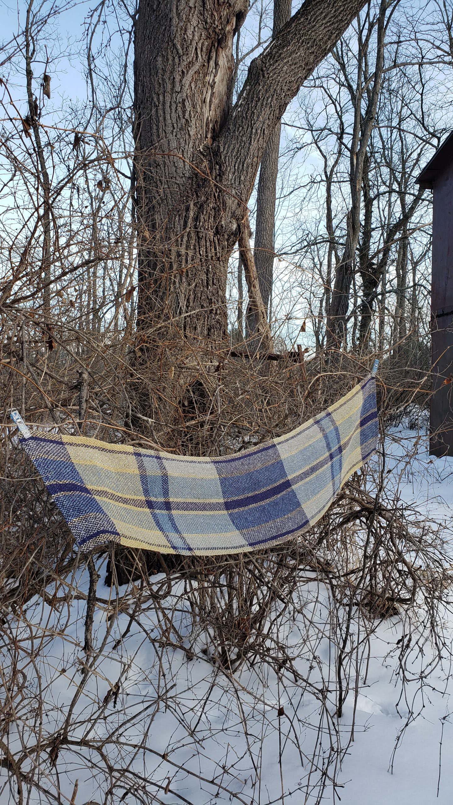 Plaid handwoven table runner draped over branches in a snowy landscape with bare trees.