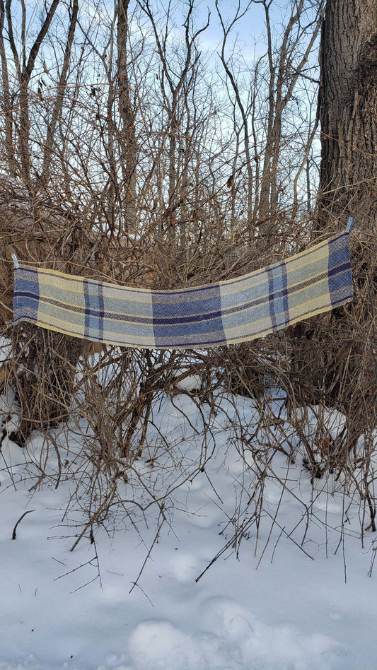 Handwoven plaid table runner draped across branches in a snowy wooded background.