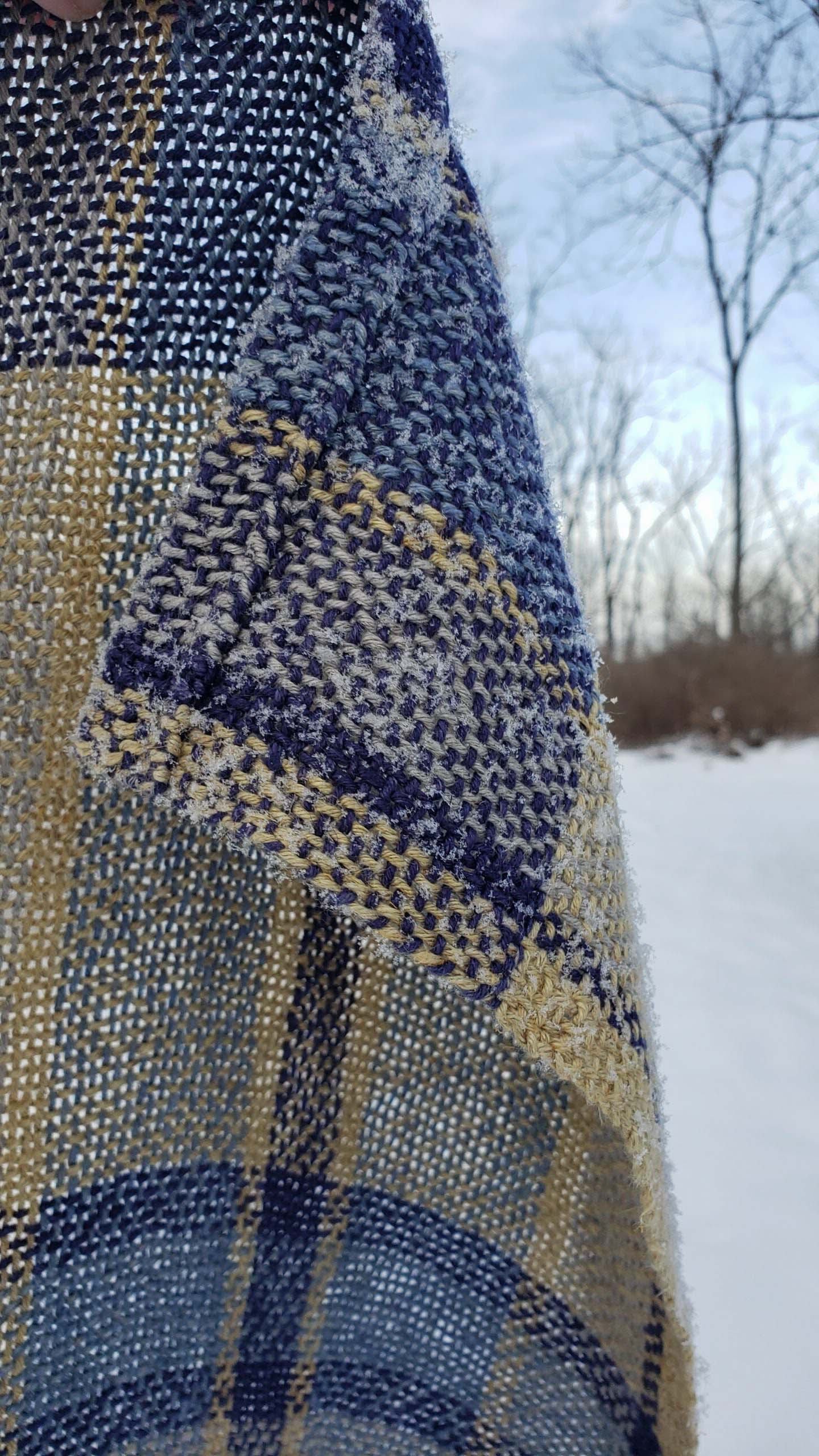 Close-up of a handwoven blue and yellow plaid fabric with a blurred snowy landscape in the background.