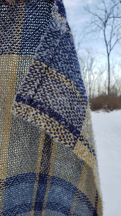 Close-up of a handwoven blue and yellow plaid fabric with a blurred snowy landscape in the background.