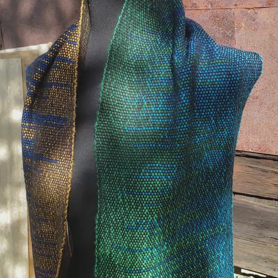 A black mannequin is wearing a handwoven clasped weft scarf in gradient jewel tones with black fringe against the outside wall of a rustic metal sided wooden building in an autumnal rural setting. The same scarf draped over a long stick shuttle suspended from an old black walnut tree. A montage of still photos of the scarf weaving process.
