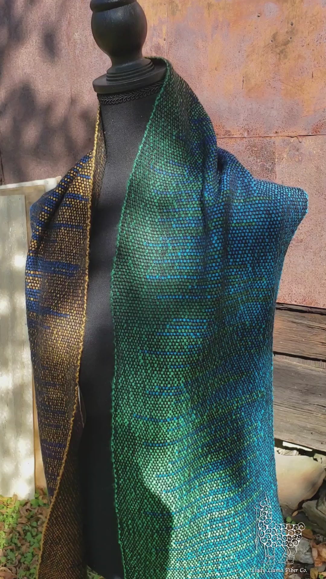 A black mannequin is wearing a handwoven clasped weft scarf in gradient jewel tones with black fringe against the outside wall of a rustic metal sided wooden building in an autumnal rural setting. The same scarf draped over a long stick shuttle suspended from an old black walnut tree. A montage of still photos of the scarf weaving process.