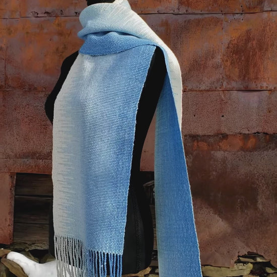 Blue and white handwoven scarf on a mannequin against a rustic exterior wall with a layer of snow on the ground.