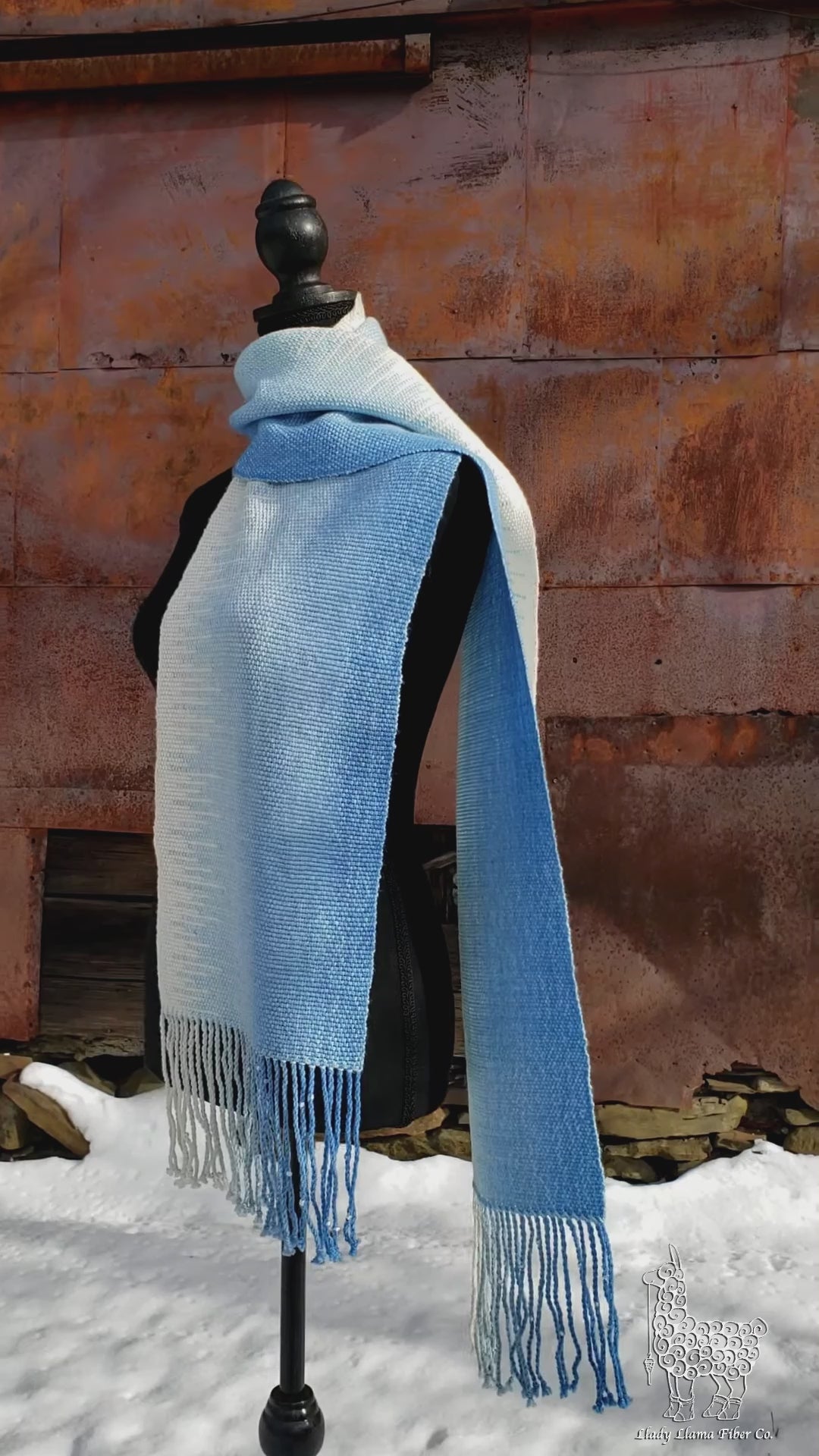 Blue and white handwoven scarf on a mannequin against a rustic exterior wall with a layer of snow on the ground.