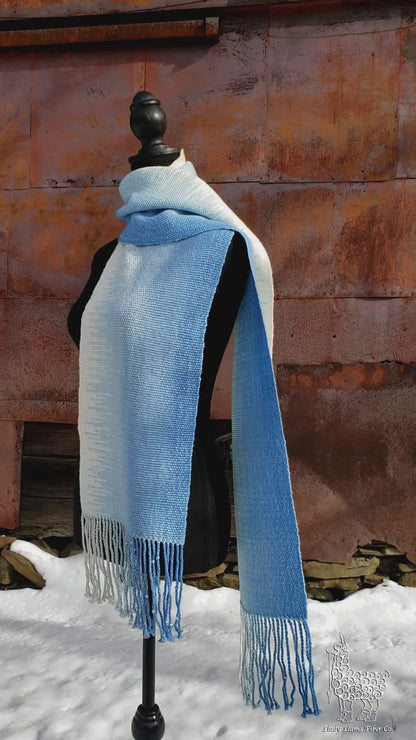 Blue and white handwoven scarf on a mannequin against a rustic exterior wall with a layer of snow on the ground.